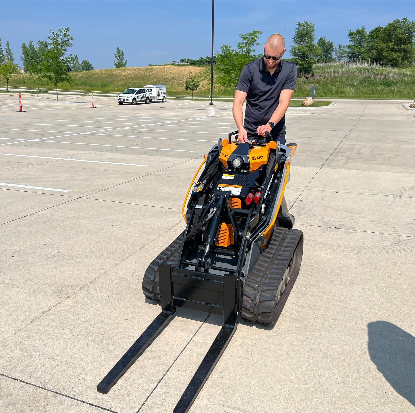 Person operating a small construction vehicle on a concrete surface with trees and vehicles in the background.